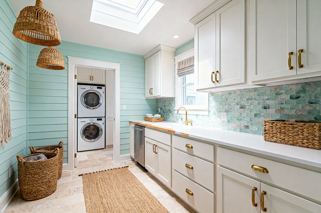 A bright coastal laundry room with seafoam shiplap walls, white shaker cabinets with brass hardware, and white quartz counters. A skylight and window illuminate an undermount sink with a brass faucet, an aqua and sage glass mosaic backsplash, and a travertine floor with a jute runner. Rattan pendant lights, woven baskets, and a stacked front-load washer and dryer in a cased niche complete the space.