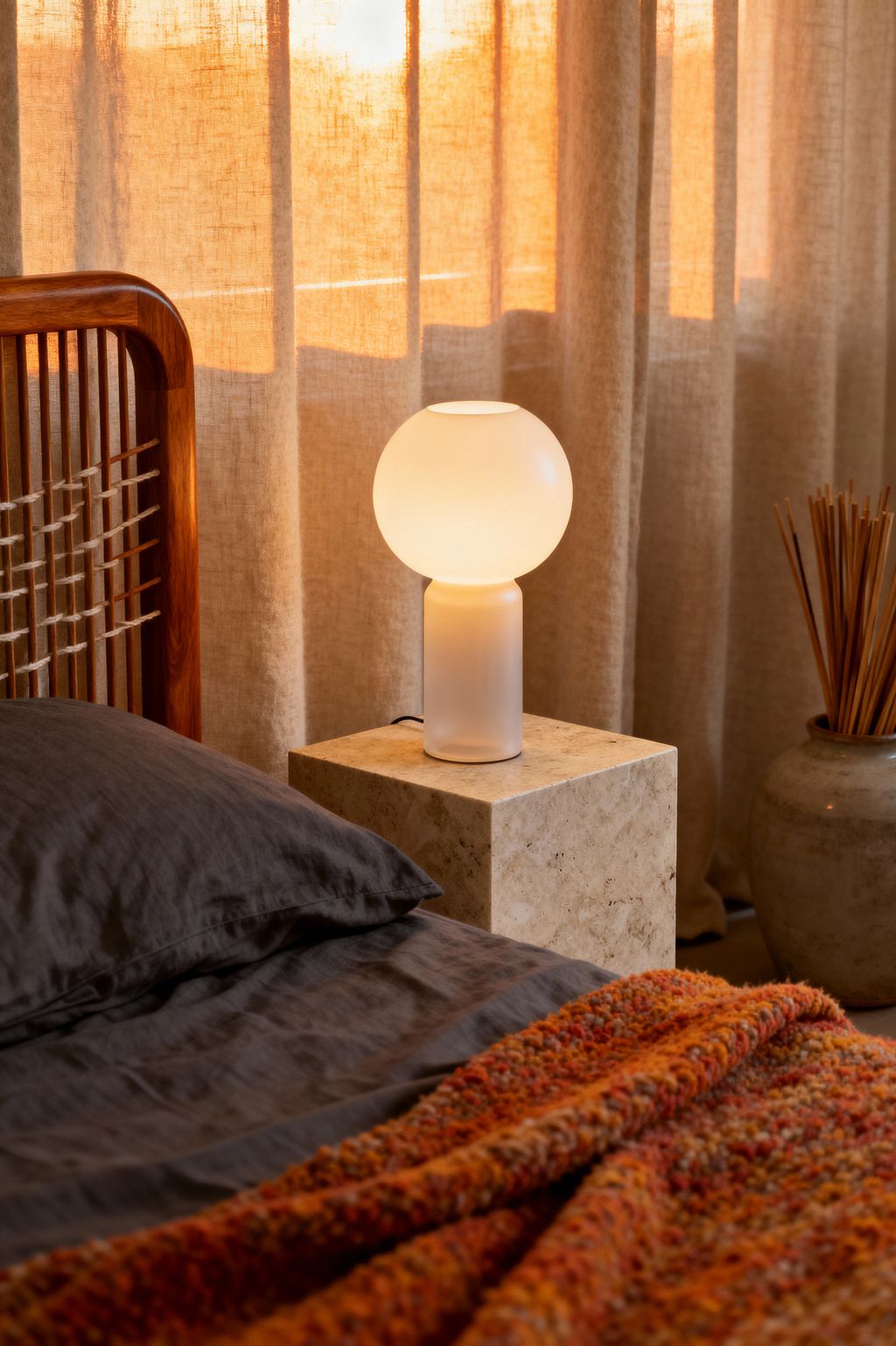 Close-up of a Scandinavian bedroom corner showing a warm-toned wooden bed with rope-laced slats, charcoal linen bedding, and a rust, chunky knit throw. A frosted glass globe lamp glows atop a travertine cube nightstand beside floor-length natural linen curtains lit by sunset, with a gray ceramic vessel of reed sticks nearby, emphasizing simple lines, tactile textures, and soft ambient warmth.