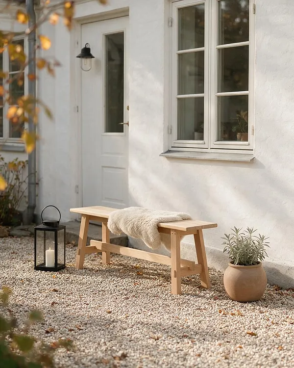A serene Scandinavian-style exterior with a white stucco wall, cream-trimmed windows, and a pale wooden bench set on a gravel courtyard. A black lantern with a candle and a terracotta plant pot add simple, natural accents.