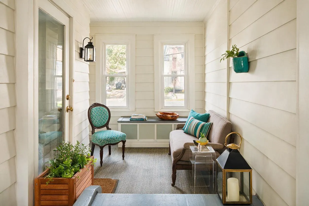 A narrow front porch with white shiplap walls, a glass-paneled door, and two tall windows at the back. The space includes a gray sofa with striped and teal pillows, a turquoise patterned chair, a built-in bench with books and a bowl, a potted plant in a wooden box, and black-and-brass lantern decor.