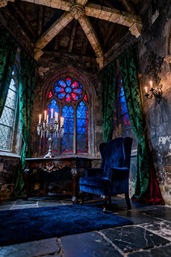 A moody Gothic living room with ribbed stone vaulting and weathered walls features pointed-arch stained-glass windows in cobalt blue and crimson, framed by emerald damask curtains with red lining. A royal-blue velvet wingback chair sits beside a carved dark-wood table topped with a silver candelabra and crystal drops. A wrought-iron wall sconce holds candles on the right, and a dark flagstone floor is softened by a deep blue rug, all illuminated by warm candlelight.