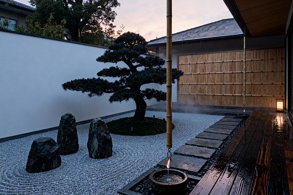 A serene Japanese Zen patio centers on a carefully pruned pine on a moss mound, encircled by raked white gravel and three dark standing stones. A bamboo rain chain drips into a small stone basin beside a wet, dark-stained wood deck, with granite stepping stones, a smooth white plaster wall, and a honey-colored bamboo screen; a soft lantern glows as evening mist gathers, creating a calm, contemplative mood.
