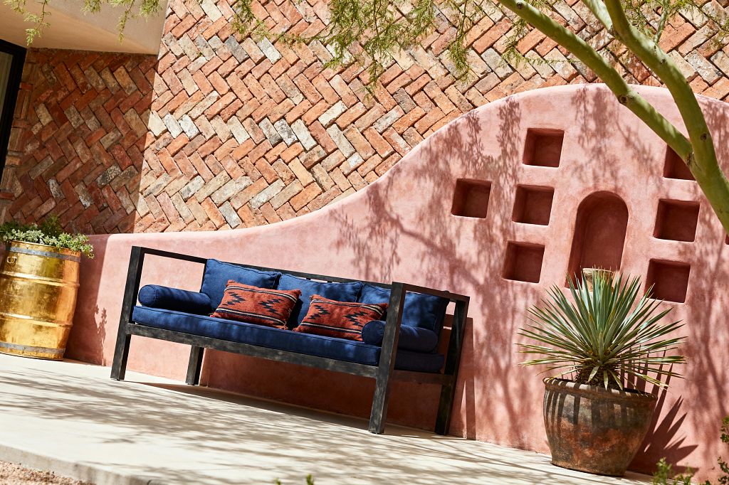 Sunlit Southwestern patio featuring a curved pink adobe stucco wall with square and arched niches in relief. In front sits a navy outdoor bench with a black metal frame, layered with indigo cushions and rust kilim pillows. Behind, a herringbone clay brick wall adds texture. A patinated brass barrel planter with succulents sits left, and a terracotta pot with a spiky agave sits right, on a pale concrete floor dappled by tree shadows.