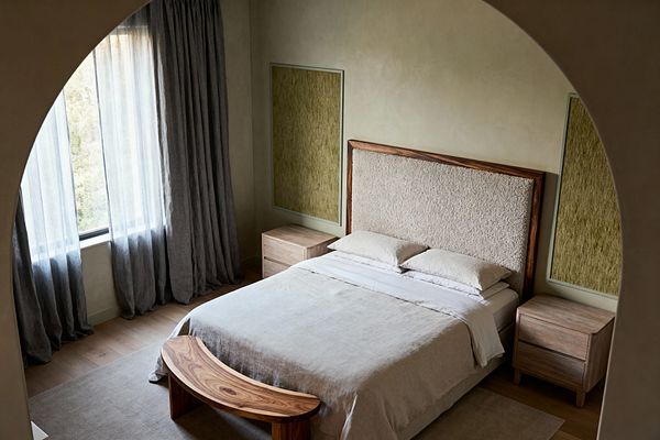 A transitional bedroom viewed through an arched opening features a bed with a tall bouclé-upholstered headboard framed in walnut and stone-colored linen bedding. Light oak flooring and a neutral rug support pale wood nightstands and a curved walnut bench, while textured olive panels flank the headboard; greige plaster walls and a large window with sheer and charcoal linen curtains diffuse soft daylight.