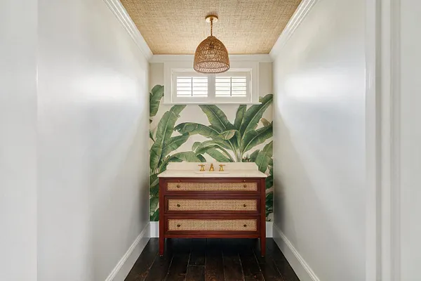 A narrow powder room with white walls and a dark wood floor features a burgundy vanity with cane-front drawers and a light countertop. The back wall is covered in bold tropical palm wallpaper, topped by a small shuttered window and a woven pendant light hanging from a woven-look ceiling.