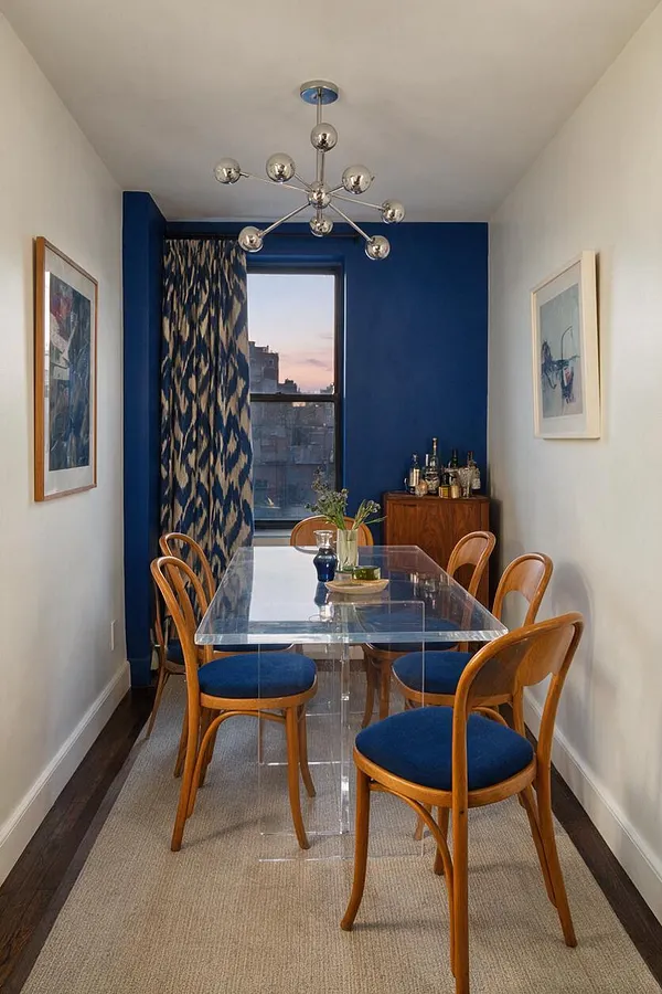 A narrow dining room features a clear acrylic table surrounded by curved wood chairs with deep blue seats, set on a light woven rug. A vivid cobalt blue back wall frames a window with patterned curtains, while white side walls display framed art and a chrome sputnik chandelier hangs overhead.