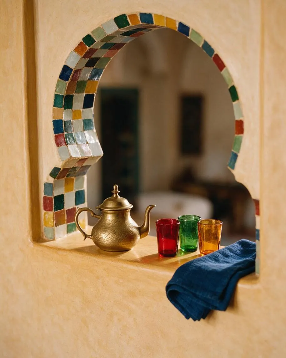 A Moroccan-style stucco niche with a rounded horseshoe arch edged in multicolored mosaic tiles. On the ledge sit a small brass teapot, three colored tea glasses in red, green, and amber, and a folded blue cloth.