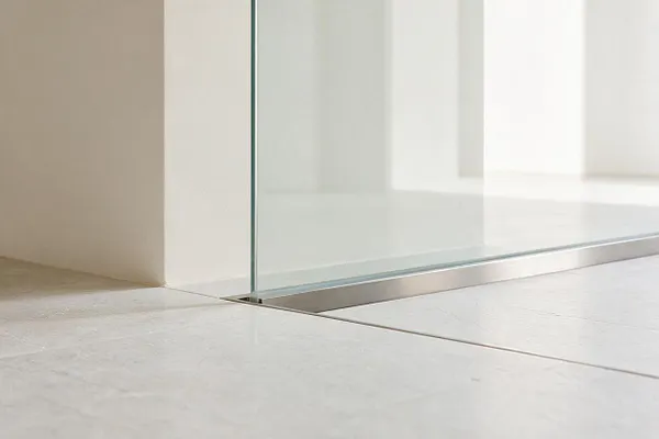 A close-up of a minimalist interior detail showing a clear glass partition set into a slim metal track beside smooth light stone or tile flooring. Soft white walls and bright natural light create a clean, airy look.