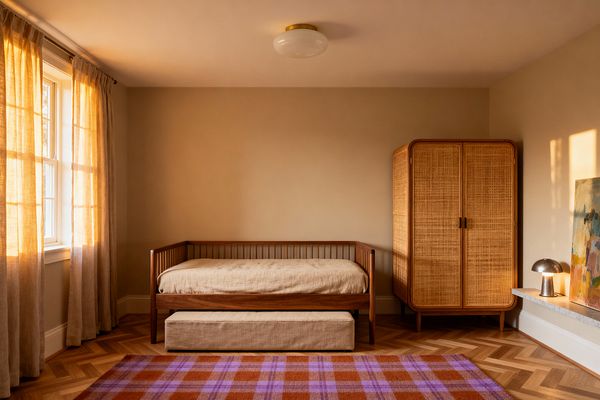 A warm, minimalist kids’ room features a walnut-toned daybed with linen bedding and a pull-out trundle beneath. At right, a rounded wood wardrobe with woven cane doors stands against taupe walls. Sunlight streams through beige sheer curtains onto a herringbone wood floor, where a terracotta and lilac plaid rug adds color. A low stone ledge holds an abstract painting and a small brushed-metal lamp, while a brass-and-opal glass ceiling light centers the space.