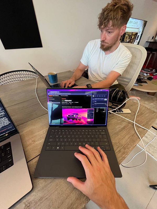 A man sits at a table working on a laptop while another laptop in the foreground is displaying the Clipbasket plugin interface in a browser window.