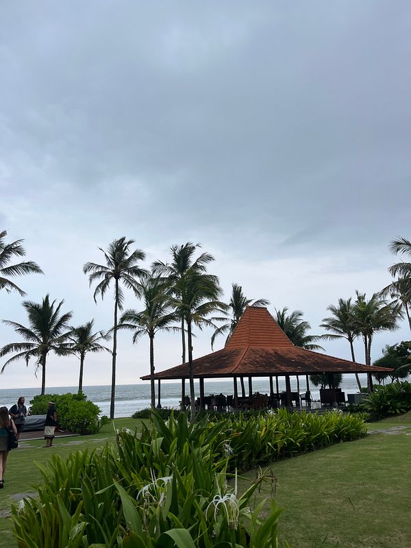A serene beachside scene featuring a traditional pavilion surrounded by palm trees and lush greenery.