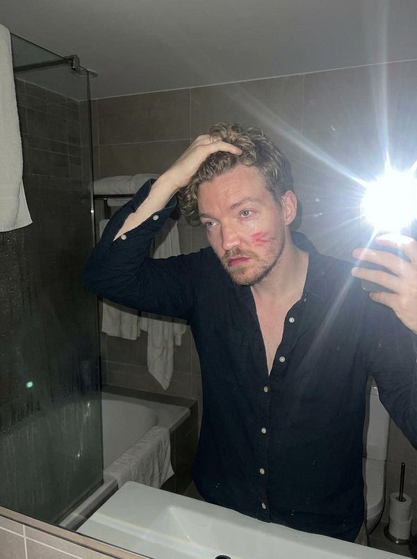 A man is taking a selfie in a bathroom mirror, showing a playful expression and a kiss mark on his cheek.