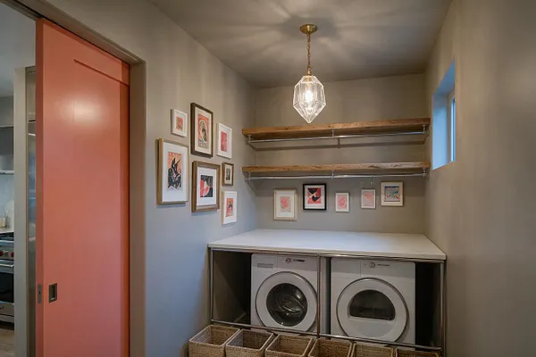 A compact eclectic laundry room with a coral sliding door, neutral walls, and a white countertop over front-loading washer and dryer. Framed artwork decorates the walls, two rustic wood shelves run above the counter, and a faceted glass pendant light hangs from the ceiling.