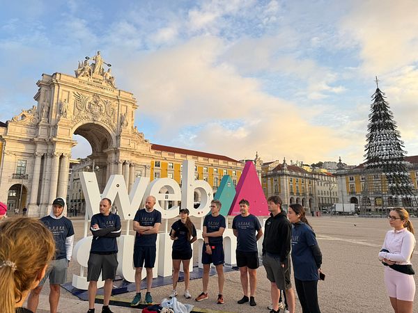 A group of individuals in matching shirts gathers for a run in front of a historic arch and a large Web Summit sign.