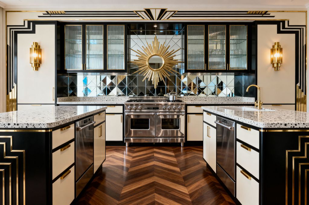 A glamorous Art Deco kitchen with two parallel islands in black and cream, each topped with speckled terrazzo and fitted with brass pulls. Centered on a stainless range, the back wall features diamond-pattern mirrored tiles and a gold sunburst, fluted-glass upper cabinets, tiered brass sconces, and bold stepped black-and-gold trim, all grounded by a warm walnut chevron wood floor in a perfectly symmetrical layout.