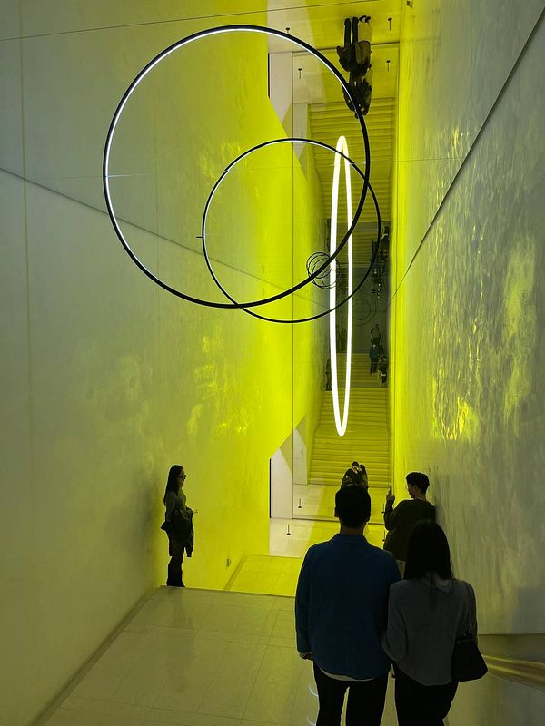 A modern staircase illuminated by vibrant yellow lighting and circular light fixtures, with visitors exploring the space.