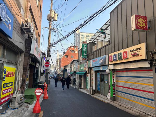 A street scene in a Seoul neighborhood with Soviet-era architectural influences, featuring a person walking and a mix of old and modern buildings.