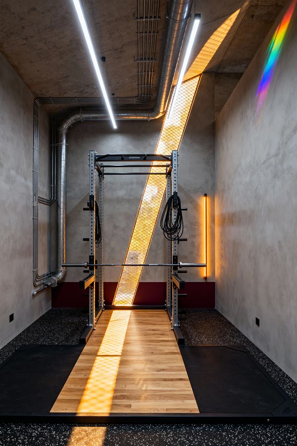 A compact industrial home gym features a steel power rack centered on an oak lifting platform set within speckled black rubber flooring. Raw concrete-like walls and ceiling expose galvanized ducts and surface-mounted conduits, while a diagonal backlit perforated metal panel and a vertical amber tube light contrast with cool white linear LEDs overhead; a narrow burgundy band runs along the rear wall.