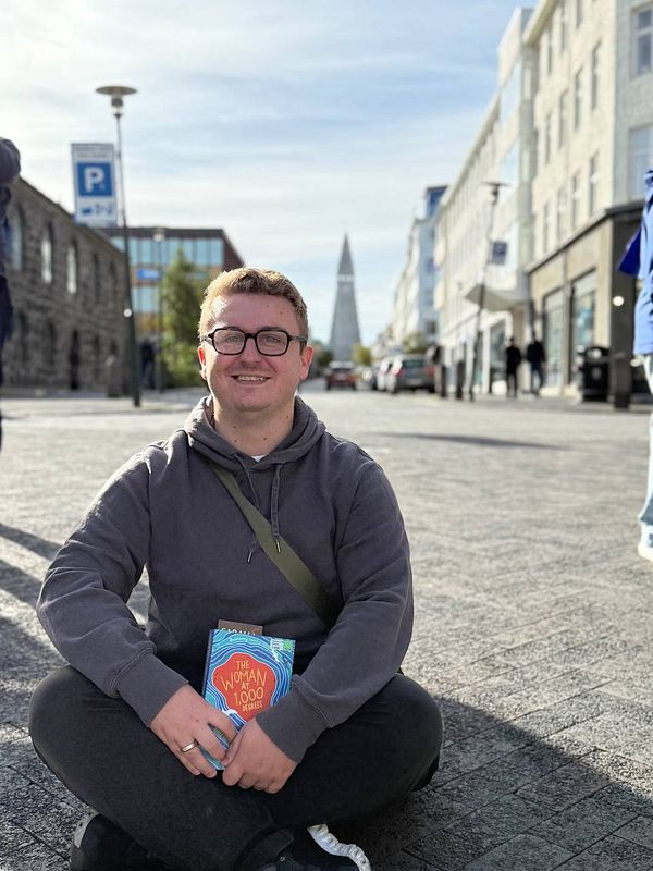 A young man sits on the pavement in Reykjavik, holding a book with a prominent church spire in the background.