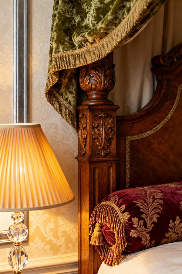 Close-up of a traditional bedroom showing a carved wooden bedpost with acanthus motifs beside a headboard trimmed with gold braid. An olive green velvet canopy drape with bullion fringe hangs above a burgundy velvet bolster with gold damask and tassels, while a crystal-and-brass table lamp with a pleated shade glows against cream damask wallpaper and a slim metallic frame on the wall.