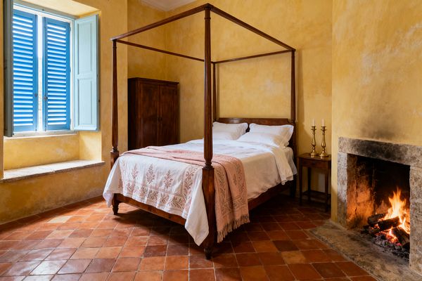 A cozy French Country bedroom features a dark-stained four-poster bed dressed in white linens and a blush embroidered throw. Limewashed ochre walls, terracotta tile floors, a stone fireplace with a live fire, a simple wood armoire, a narrow bedside table with brass candlesticks, and sky-blue interior shutters over a deep stone window seat create a warm, rustic atmosphere.