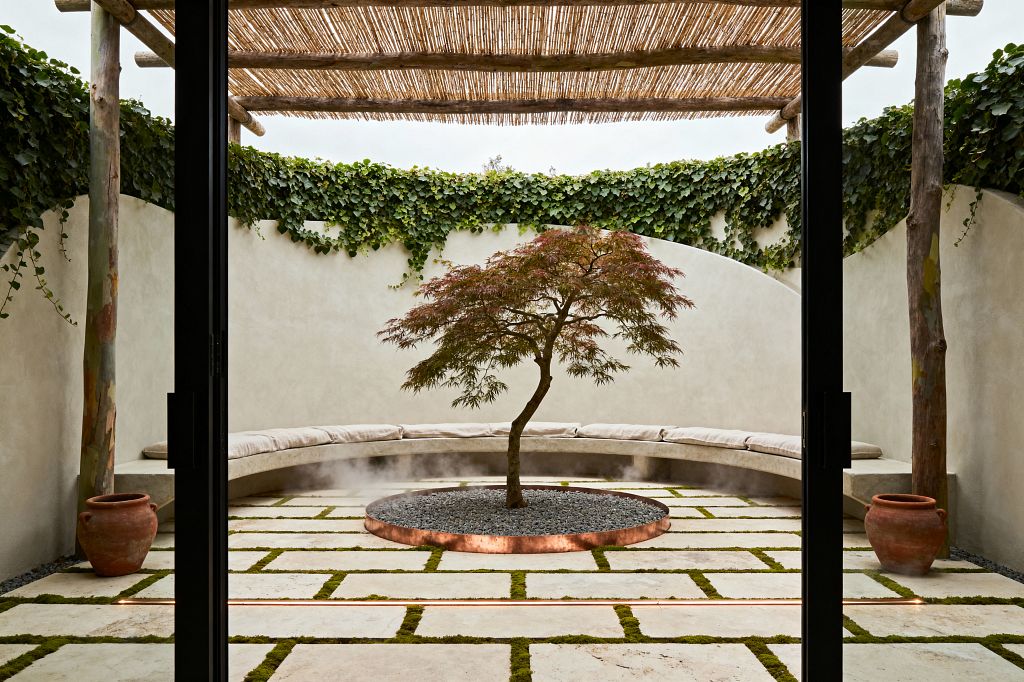 An enclosed patio with curved sand-colored stucco walls and a rustic pergola of timber posts and reed slats. At the center, a small Japanese maple rises from a copper-edged circle of black pebbles with soft mist. The floor is a grid of pale limestone pavers with green moss joints and a subtle linear light. A built-in curved bench with light beige cushions lines the wall, flanked by terracotta pots and trailing ivy.