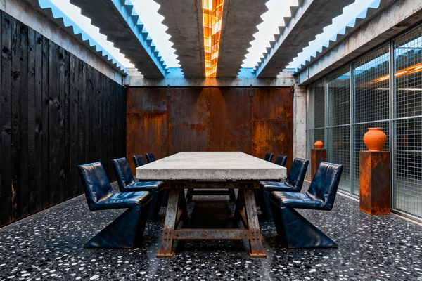 A long concrete dining table on rusted steel trestles stands on a black terrazzo floor, surrounded by eight deep-blue cantilever chairs. The room features a charred wood plank wall on one side, a backdrop of patinated steel panels, and floor-to-ceiling wire-mesh glazing; above, exposed sawtooth concrete beams and a central amber-lit skylight illuminate rusted pedestals topped with terracotta pots.