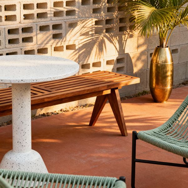An outdoor patio features a round white speckled terrazzo pedestal table beside a slatted teak bench set in front of a concrete breeze-block wall. A sage green woven-rope chair with a black metal frame and a tall brushed brass planter holding a palm sit on terracotta-colored concrete, with warm afternoon light casting the palm’s shadows across the wall.