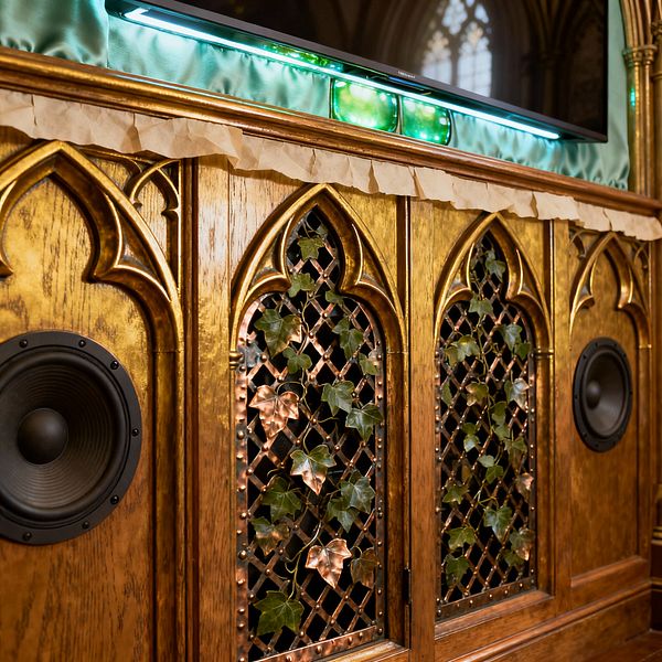 A Victorian Gothic media console features carved pointed arches in burnished golden wood, with copper lattice grilles adorned by trailing ivy between integrated black speakers. Above, a flat-screen TV is set against ruched pale-green satin and illuminated by teal LED lighting, while a crinkled parchment-like trim softens the edge, creating an ornate yet contemporary media focal point.