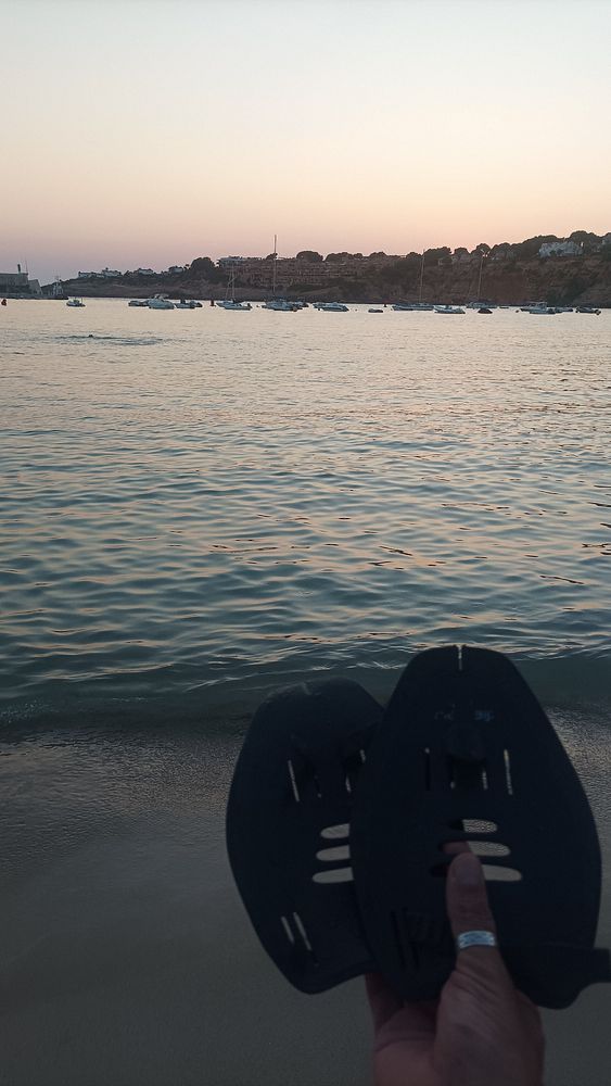 A person holds swim fins while standing on a beach at sunset, with boats in the water and a scenic coastline in the background.