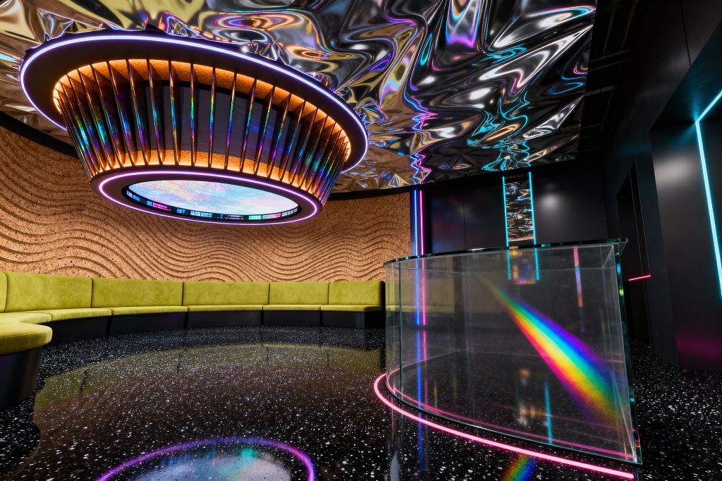 A futuristic media room with a curved lime-green banquette set against a rippled cork wall. Overhead, a mirrored liquid-like ceiling reflects an oversized circular halo fixture with iridescent fins and a central digital display. The high-gloss black speckled floor mirrors neon magenta and cyan light. At right, a curved clear-glass console glows with perimeter LEDs and shows a rainbow prism, while black wall panels feature vertical light strips.