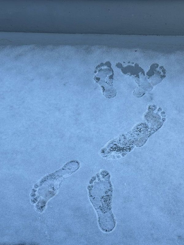 A child in a winter jacket and hat standing barefoot in the snow with a joyful expression.