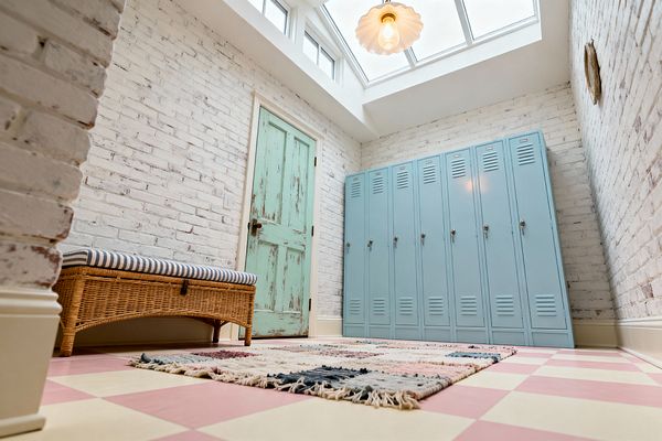 A light-filled mudroom with whitewashed brick walls features a row of powder-blue metal lockers on the right and a distressed mint wooden door at the far end. A rattan bench with a blue-and-white striped cushion sits on a pink and buttercream checkerboard floor layered with a fringed flat-weave rug, while a skylight and scalloped glass pendant provide warm illumination.