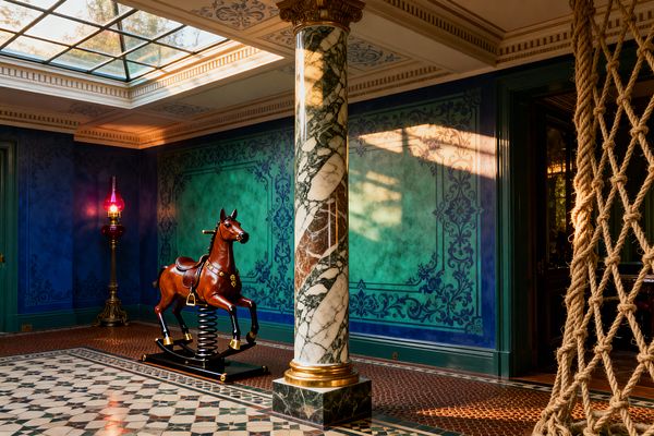 A sunlit home gym with a glazed ceiling skylight features a veined green-and-white marble column with a gold base, ombré teal walls framed by navy stenciling, and patterned ceramic mosaic flooring with a terracotta border. A glossy mahogany spring rocking horse, a thick sisal rope climbing net, green-painted doors and trim, and a brass floor lamp with a pink blown-glass shade complete the richly detailed Victorian setting.