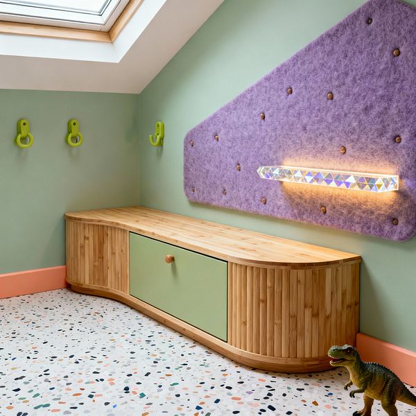 A contemporary kids’ corner beneath a skylight shows mint green walls, a curved low bench in light bamboo with a sage cabinet front, and a lilac wool-felt pinboard dotted with wooden pegs. An iridescent linear light glows from the panel above terrazzo flooring with multicolored chips and a peach baseboard, with three lime wall hooks and a small toy dinosaur on the floor.