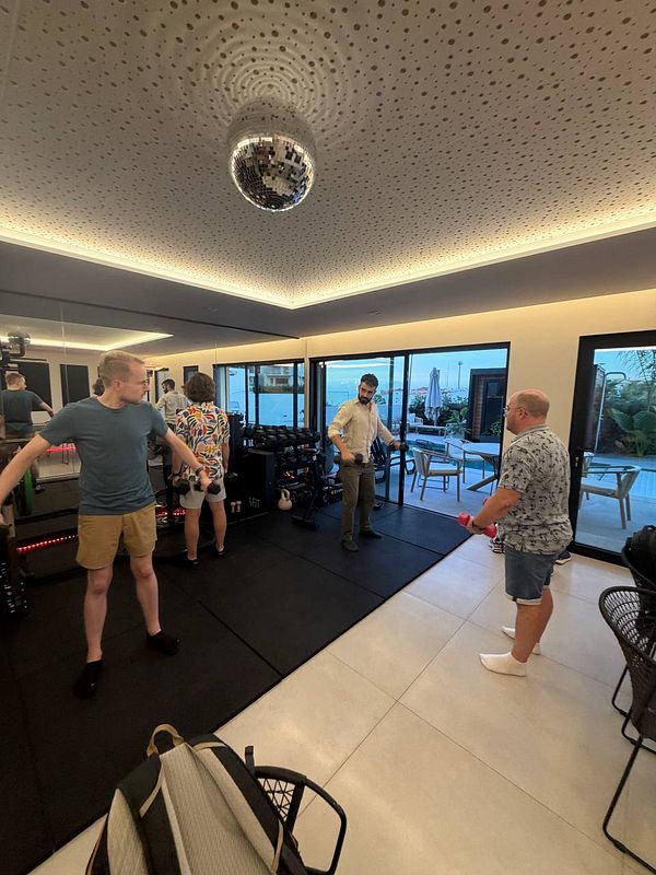 Four men exercising with dumbbells in a modern room with black mats and a disco ball on the ceiling.