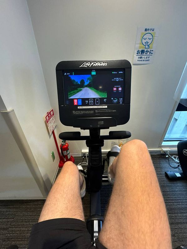 A person is using a stationary bike in a gym with a digital display showing a scenic route.