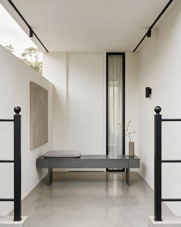 A minimalist front porch with white walls, a smooth concrete floor, and black metal railings and track lighting. A dark gray bench with a cushion sits against the back wall beside a tall narrow window, creating a calm, spare entry space.