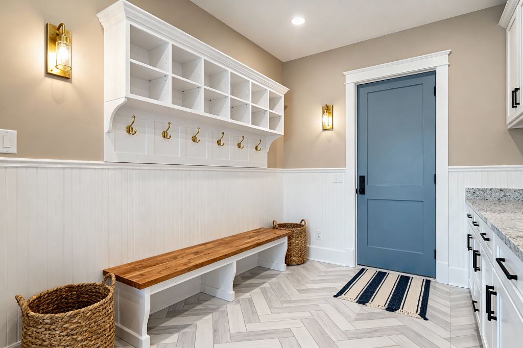 A bright mudroom with white beadboard wainscoting and built-in open cubbies above a long wood bench fitted with brass coat hooks. A slate-blue door with black hardware faces white shaker cabinetry with a light speckled stone countertop; light gray herringbone tile flooring, woven baskets, a navy-and-cream striped rug, recessed lighting, and brass-and-glass wall sconces complete the organized, transitional entry.