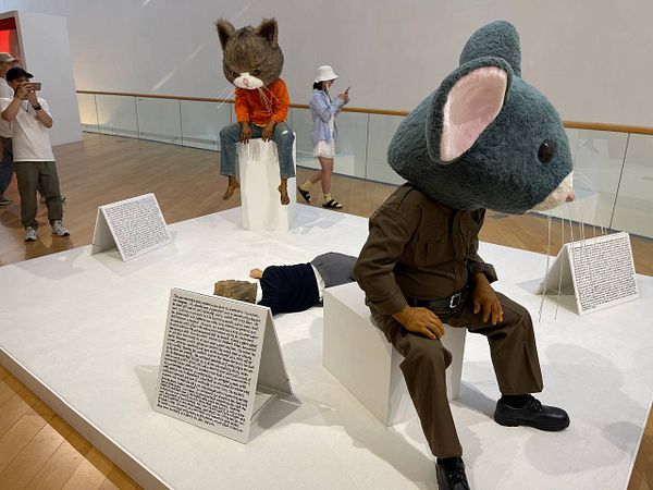 The image features a contemporary art installation with anthropomorphic sculptures and visitors observing the artwork.