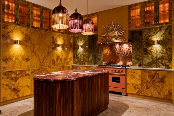 A dramatic maximalist kitchen wrapped in golden, heavily veined onyx-style cabinetry with ribbed amber glass uppers centers on a rounded island with a fluted walnut base and a deep red marble countertop. A hammered copper backsplash and hood with a sunburst accent frame a burnt-orange range, while brass sconces and three plum velvet fringe pendants cast warm light over the richly patterned surfaces and the pale stone floor.