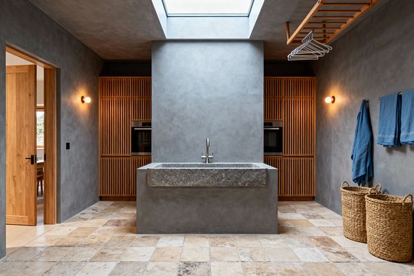 A Scandinavian-style laundry room features smooth gray microcement walls and a large honed granite utility sink on a matching plinth beneath a skylight. Slatted oak cabinetry flanks a central column and houses two integrated black-glass appliances. The floor is tumbled limestone in warm beige tones. Brass sconces, a ceiling-mounted wooden drying rack with white hangers, blue towels on a rail, and two woven baskets complete the calm, functional space.
