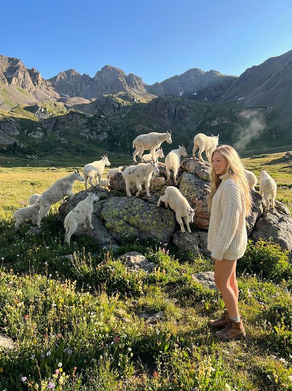 A young woman stands smiling among a group of mountain goats in a scenic mountainous landscape.