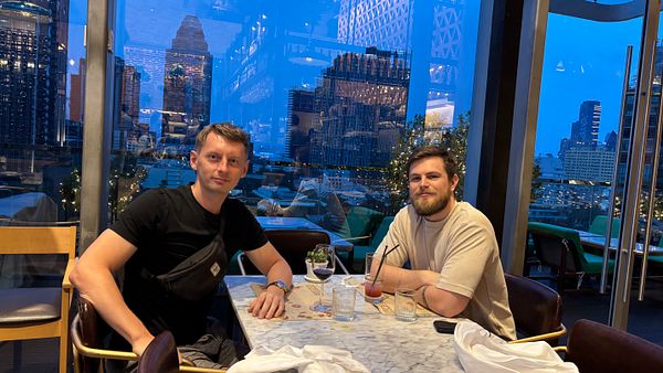 Two men are seated at a table in a restaurant with a city skyline view in Bangkok.