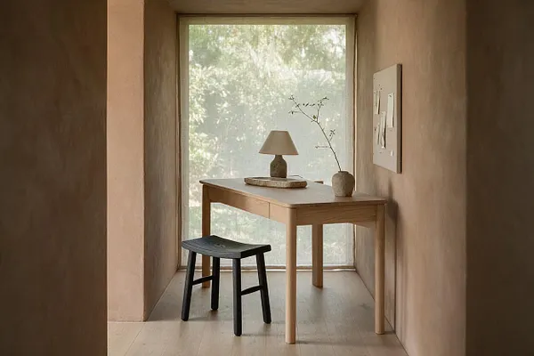 A narrow home office with a light oak desk placed in front of a large window, paired with a small black stool. A simple table lamp and a slender branch in a vase sit on the desk, while warm taupe walls and pale flooring create a calm, minimalist atmosphere.