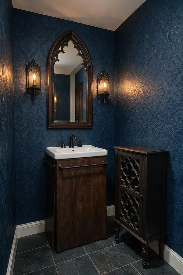 A compact powder room with deep blue patterned walls, a pointed Gothic-style arched mirror, and two warm wall sconces. A white sink sits on a dark wood vanity, with a matching carved storage cabinet and slate-look floor tiles below.