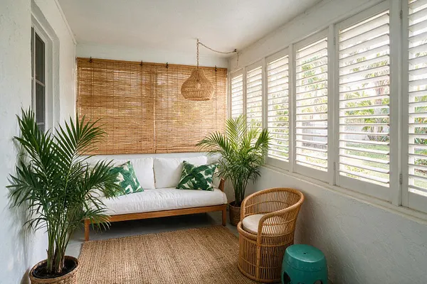 A bright tropical backyard lounge with a white sofa, woven bamboo blinds, louvered windows, and multiple potted palms. A wicker chair, jute rug, and woven pendant light add natural texture to the relaxed space.