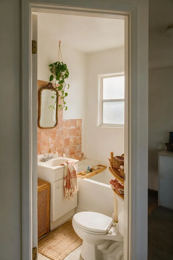 A small master bathroom with a white bathtub, toilet, and compact vanity styled in a warm bohemian look. The space features terracotta wall tiles, a vintage gold-framed mirror, a hanging pothos plant, woven baskets, a jute rug, and soft neutral walls with natural light from a frosted window.