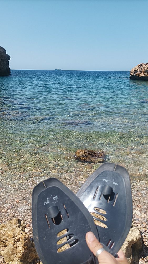 A person holds a pair of swimming fins in front of a clear blue sea.