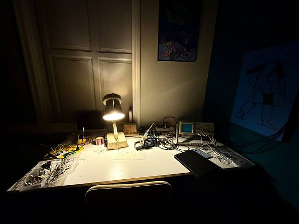 A cluttered work desk illuminated by a lamp, featuring various electronic components and tools.